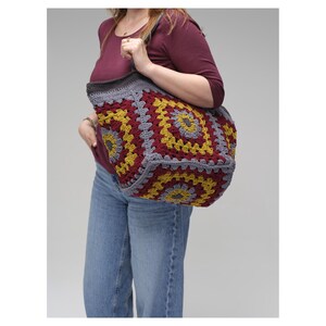 May include: A large, handmade crochet tote bag constructed from multiple square motifs. The bag features a repeating pattern of deep burgundy, mustard yellow, and grey yarn, finished with a solid grey border and two sturdy shoulder straps.
