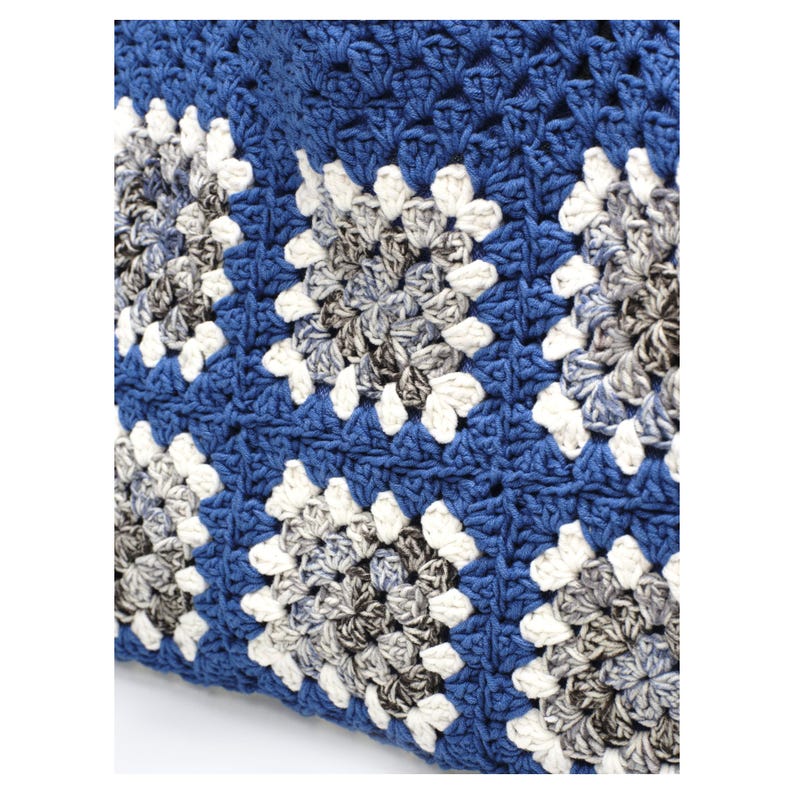 Puede incluir: Un tejido de ganchillo con un patr&oacute;n repetitivo de cuadrados granny. Los cuadrados est&aacute;n construidos con un hilo central gris y blanco jaspeado, bordeados por un hilo color crema s&oacute;lido y unidos con un hilo azul profundo s&oacute;lido.