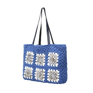 Puede incluir: Un bolso tote de ganchillo azul con un patr&oacute;n de seis motivos cuadrados. Cada cuadrado tiene un borde blanco que rodea un centro de hilo gris y blanco matizado. El bolso incluye dos correas largas de hombro azul oscuro tipo cuero.