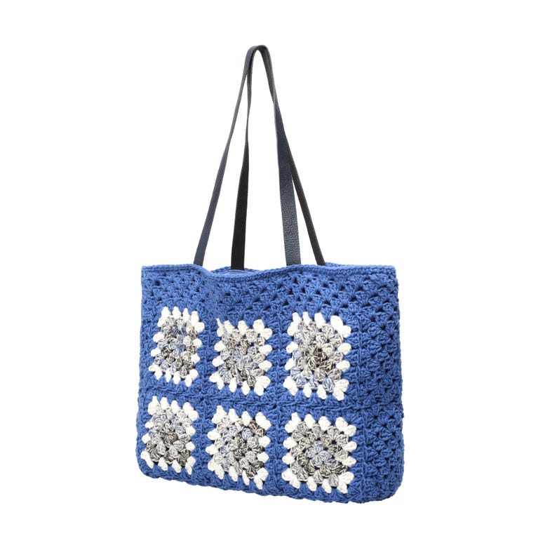 Puede incluir: Un bolso tote de ganchillo azul con un patr&oacute;n de seis motivos cuadrados. Cada cuadrado tiene un borde blanco que rodea un centro de hilo gris y blanco matizado. El bolso incluye dos correas largas de hombro azul oscuro tipo cuero.