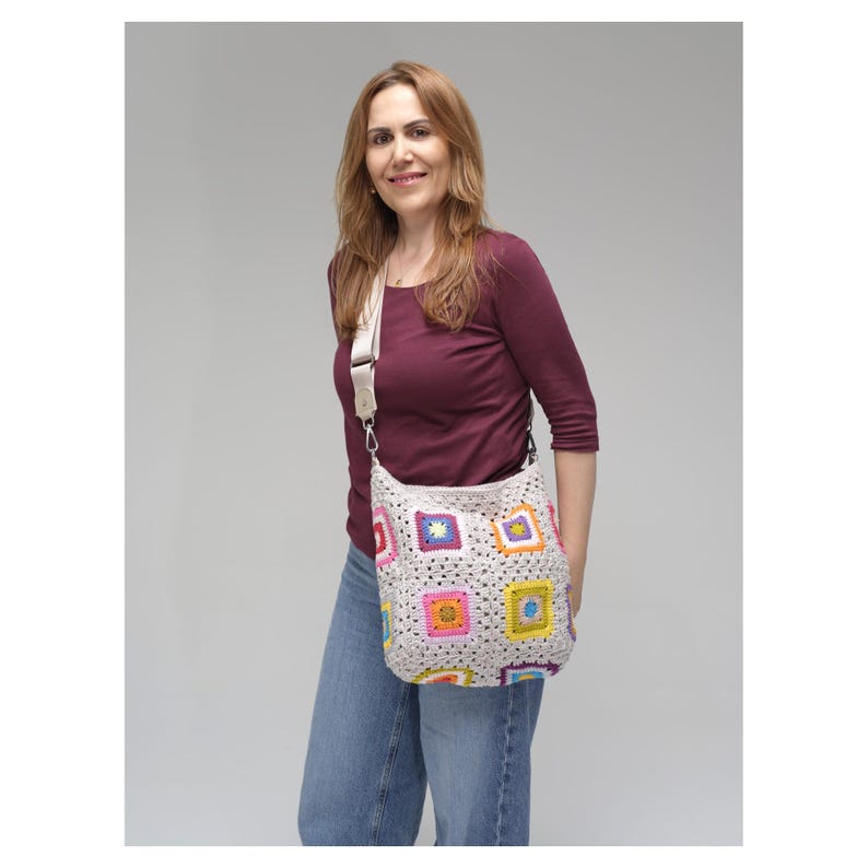 May include: A cream-coloured crochet shoulder bag with colourful square patterns. The bag has a long, adjustable strap and a rounded shape. The squares feature bright colours like pink, yellow, blue, and green.