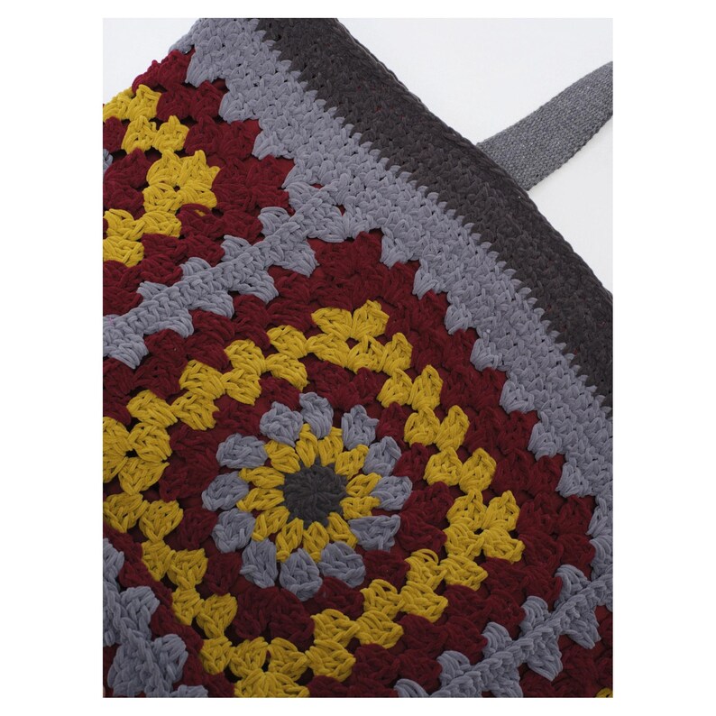 May include: A crochet tote bag featuring a square granny motif pattern in shades of mustard yellow, deep burgundy, and light gray. The bag is constructed with a dark gray solid crochet border and includes a matching dark gray fabric strap.