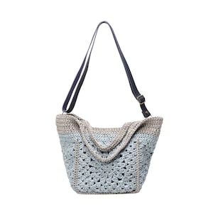 May include: A crocheted tote bag with a light blue and beige color scheme. The bag features a geometric pattern and a dark blue shoulder strap with a brass buckle. The bag is designed with a wide opening and a sturdy construction.