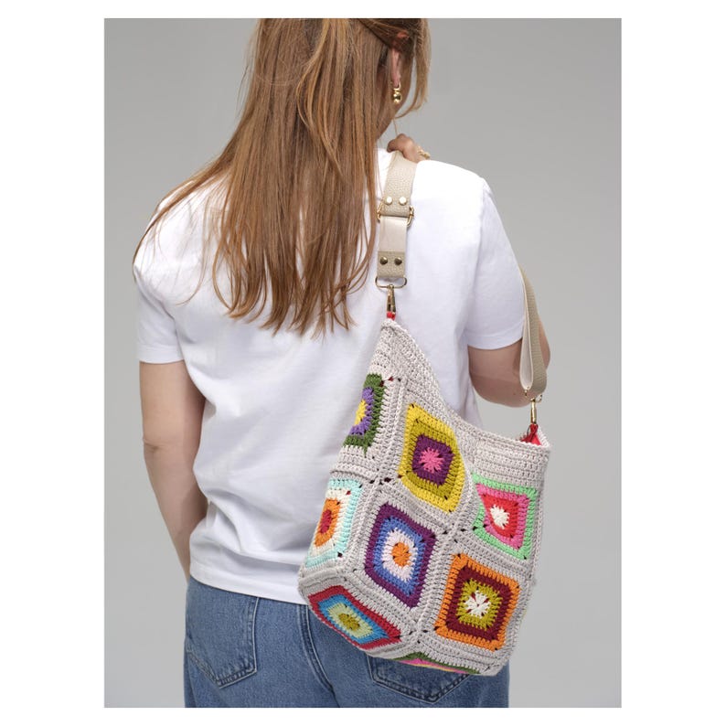 K&ouml;nnte beinhalten: Geh&auml;kelte Umh&auml;ngetasche mit Patchwork-Design. Die Tasche hat bunte Quadrate in verschiedenen Farbt&ouml;nen, darunter Gelb, Gr&uuml;n, Blau und Lila, vor einem neutralen grauen Hintergrund. Sie hat einen beigen Riemen und Metallbeschl&auml;ge.