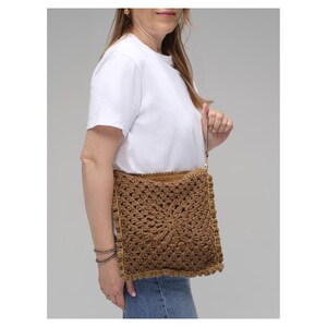 May include: A square brown crochet handbag featuring a textured granny square pattern with a central star motif. The bag has a thin wrist strap, a visible tan interior lining, and a decorative border edge, providing a compact accessory option.