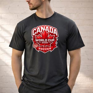 May include: Dark gray t-shirt featuring a red and white graphic. The graphic includes the word "CANADA" above a red maple leaf, with "WORLD CUP 2026" and "SOCCER" below. The shirt is short-sleeved.