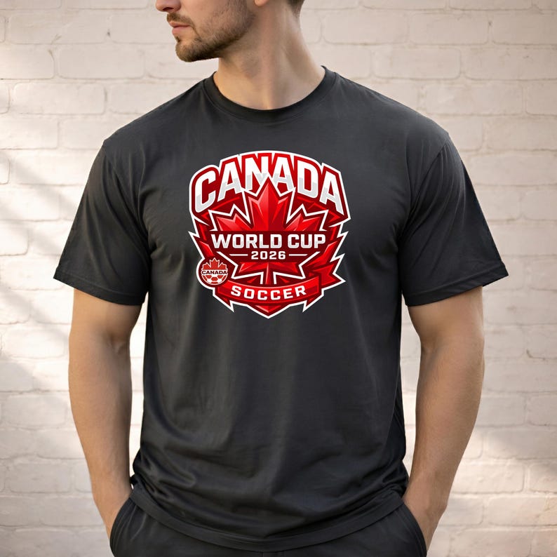 May include: Dark gray t-shirt featuring a red and white graphic. The graphic includes the word "CANADA" above a red maple leaf, with "WORLD CUP 2026" and "SOCCER" below. The shirt is short-sleeved.