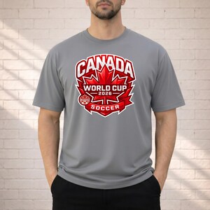 May include: A gray t-shirt featuring a red and white graphic for the Canada World Cup 2026 Soccer. The design includes a maple leaf and the text "CANADA WORLD CUP 2026 SOCCER". The shirt is short-sleeved.