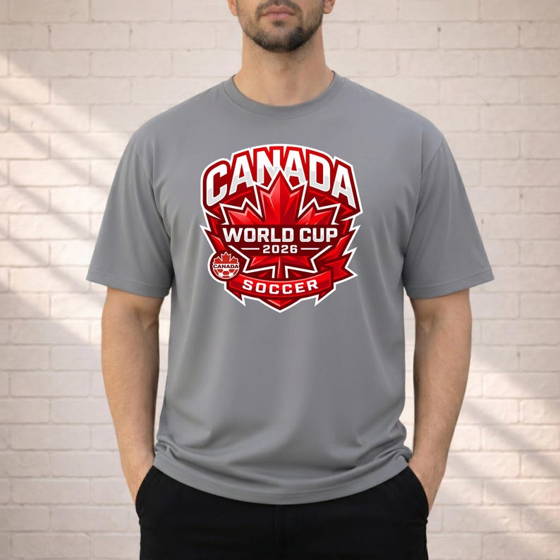 May include: A gray t-shirt featuring a red and white graphic for the Canada World Cup 2026 Soccer. The design includes a maple leaf and the text "CANADA WORLD CUP 2026 SOCCER". The shirt is short-sleeved.
