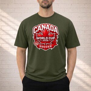 May include: Olive green t-shirt featuring a graphic design for the 2026 World Cup Soccer in Canada. The design includes the word "Canada" in white, a red maple leaf, and the words "World Cup 2026 Soccer".