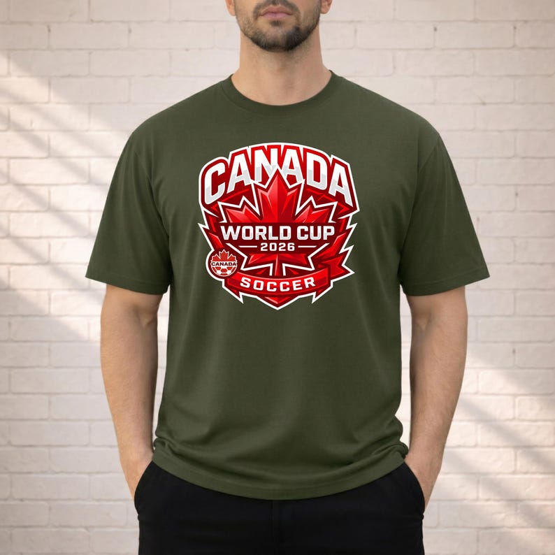 May include: Olive green t-shirt featuring a graphic design for the 2026 World Cup Soccer in Canada. The design includes the word "Canada" in white, a red maple leaf, and the words "World Cup 2026 Soccer".
