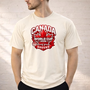 May include: Cream-colored t-shirt featuring a large red and white graphic. The graphic includes the word "CANADA" above a maple leaf design, with "WORLD CUP 2026" and "SOCCER" below. The shirt is short-sleeved.