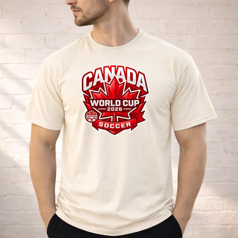 May include: Cream-colored t-shirt featuring a large red and white graphic. The graphic includes the word "CANADA" above a maple leaf design, with "WORLD CUP 2026" and "SOCCER" below. The shirt is short-sleeved.
