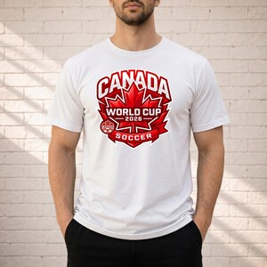 May include: White t-shirt featuring a red graphic for the 2026 Canada World Cup Soccer. The design includes a maple leaf, the word "CANADA" in bold letters, and the text "WORLD CUP 2026 SOCCER".