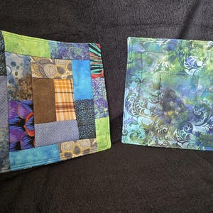 May include: Two handmade square quilted pot holders. One features a patchwork design with blue, green, brown, and purple fabrics. The other has a swirling floral pattern in shades of green, blue, and purple.