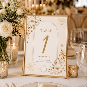 Puede incluir: Un letrero de mesa rectangular en un marco dorado con el texto TABLE 1 y THANK YOU FOR CELEBRATING WITH US. El diseño incluye un borde ovalado, motas doradas e ilustraciones florales en acuarela en tonos crema y beige.