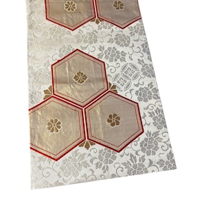 May include: A rectangular textile featuring a white background with a silver floral pattern. The fabric is decorated with five gold-toned hexagonal shapes, each outlined in red and centred with a small gold flower motif, arranged in a cluster.