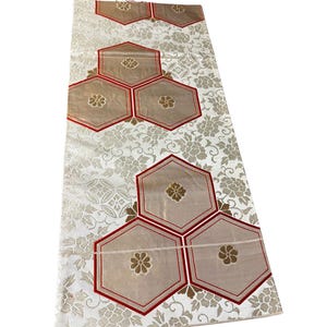 Könnte beinhalten: Ein rechteckiger Textilläufer mit weißem Hintergrund und einem sich wiederholenden Blumenmuster in Metallic-Gold. Das Design umfasst mehrere große, beigefarbene Sechsecke mit roter Umrandung, die jeweils ein goldenes Blumenemblem tragen.