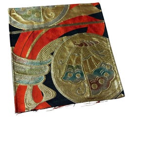 Puede incluir: Una pieza textil rectangular con fondo negro, cintas rojas vibrantes y grandes medallones dorados circulares. Los medallones contienen intrincados patrones tejidos en tonos azul, verde y dorado, creando una apariencia texturizada.