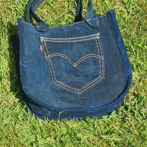Op de afbeelding: Een donkerblauwe denim tote bag met een zak en handvatten. De tas heeft een klassiek zakontwerp met beige stiksels en een rood label. De tas ligt op groen gras.