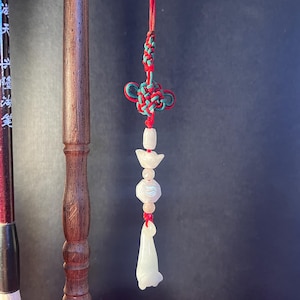 Peut inclure: Un pendentif nœud chinois décoratif avec un cordon rouge, comprenant un nœud vert et rouge, des perles blanches et roses, et un pendentif en jade blanc. Le pendentif est suspendu à côté d'un porte-pinceau en bois.