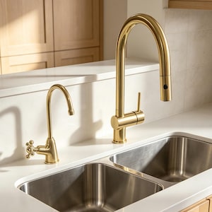 Könnte beinhalten: Ein goldfarbenes Küchenarmaturen-Set bestehend aus einer hohen, bogenförmigen Ausziehbrause mit seitlichem Hebel und einer kleineren, zusätzlichen Armatur mit Kreuzgriff, montiert auf einer weißen Arbeitsplatte über einem Edelstahl-Doppelspülbecken.