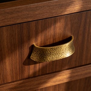 May include: A dark brown wooden drawer front featuring a horizontal, curved metal handle with a brass finish and a textured, hammered surface. The handle is mounted centrally on the drawer, which displays a visible wood grain pattern throughout.