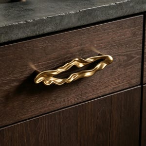 Puede incluir: Un tirador de caj&oacute;n de metal dorado con una forma org&aacute;nica, fluida y ondulada. El tirador presenta un centro hueco y una superficie lisa y reflectante, montado horizontalmente en el frente de un caj&oacute;n de madera de color marr&oacute;n oscuro.