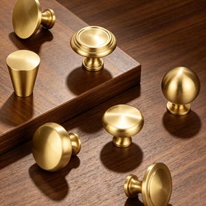 May include: A collection of seven brass cabinet knobs in various shapes, including spherical, conical, and flat-topped designs. Each knob features a smooth, brushed metallic finish with a warm gold tone, displayed in a scattered arrangement.