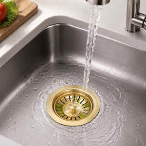 May include: A brushed gold kitchen sink drain strainer set into a stainless steel sink basin. Water flows from a silver faucet directly into the center of the circular drain, which features a series of narrow slots and a central knob for operation.