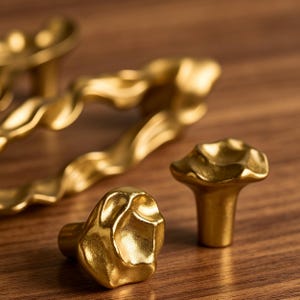 Puede incluir: Dos pomos de armario peque&ntilde;os de color dorado con una superficie irregular, org&aacute;nica y texturizada en primer plano, con un tirador de armario largo, ondulado y de color dorado a juego desenfocado al fondo.