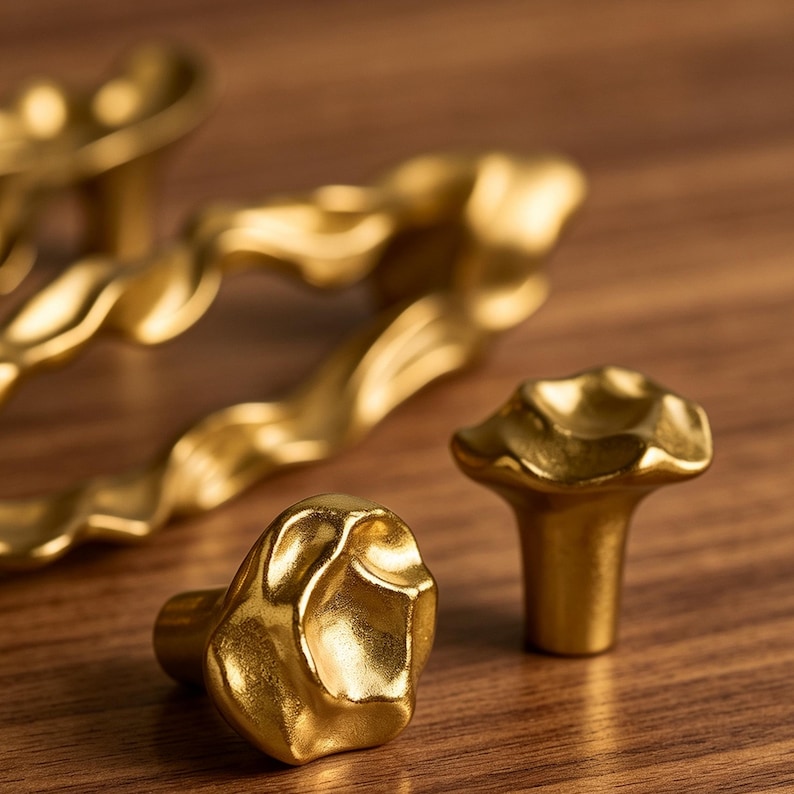 Puede incluir: Dos pomos de armario peque&ntilde;os de color dorado con una superficie irregular, org&aacute;nica y texturizada en primer plano, con un tirador de armario largo, ondulado y de color dorado a juego desenfocado al fondo.