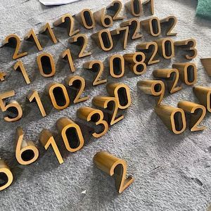 May include: A collection of three-dimensional, gold-colored metal house numbers featuring a block font. The numbers are arranged in various orientations, showcasing their depth and thickness. Each digit is individually cast with a smooth finish.