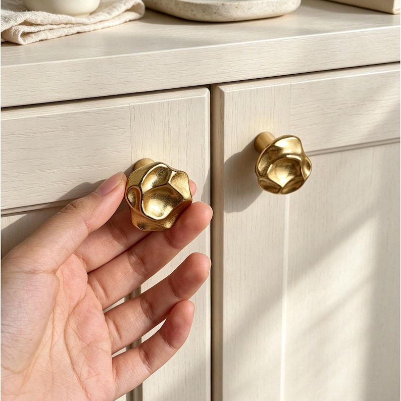 Puede incluir: Un juego de dos pomos para armario de color dorado con una superficie irregular, org&aacute;nica y texturizada. Los pomos tienen un acabado met&aacute;lico y una forma redondeada y multifac&eacute;tica. Un pomo est&aacute; fijado a una puerta de armario de madera clara.