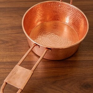 May include: A round copper pan featuring a hammered texture on the interior and exterior surfaces. The pan includes a long, flat-ended handle attached with two rivets and a small, arched loop handle on the opposite side for balanced lifting.