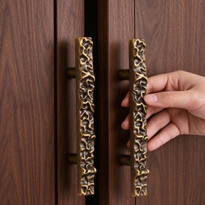 Può includere: Due maniglie verticali per armadietti in ottone anticato con una superficie ruvida e irregolare. Ogni maniglia presenta un design a barra rettangolare lunga con un motivo organico accidentato e due supporti cilindrici su un'anta in legno scuro.