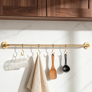 May include: A brushed gold metal wall-mounted rail featuring four matching S-hooks. Hanging from the hooks are a clear textured glass mug, a beige fabric towel, a wooden kitchen spatula, and a black round-headed kitchen ladle.