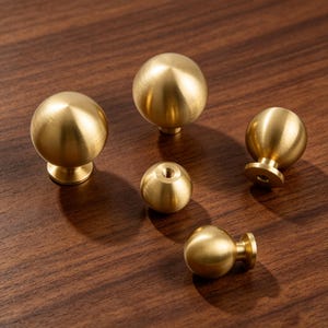 May include: A collection of five spherical cabinet knobs made of brushed brass. The knobs feature a smooth, rounded top and a small cylindrical base with a threaded hole for mounting. The finish has a soft, metallic gold-toned luster.