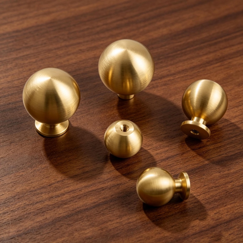 May include: A collection of five spherical cabinet knobs made of brushed brass. The knobs feature a smooth, rounded top and a small cylindrical base with a threaded hole for mounting. The finish has a soft, metallic gold-toned luster.