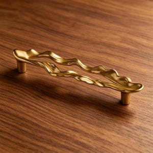 Puede incluir: Un tirador de mueble de metal dorado con una silueta larga, horizontal, ondulada y org&aacute;nica. El tirador est&aacute; sostenido por dos postes cil&iacute;ndricos en cada extremo, creando una forma continua, fluida y ondulante con un acabado suave.