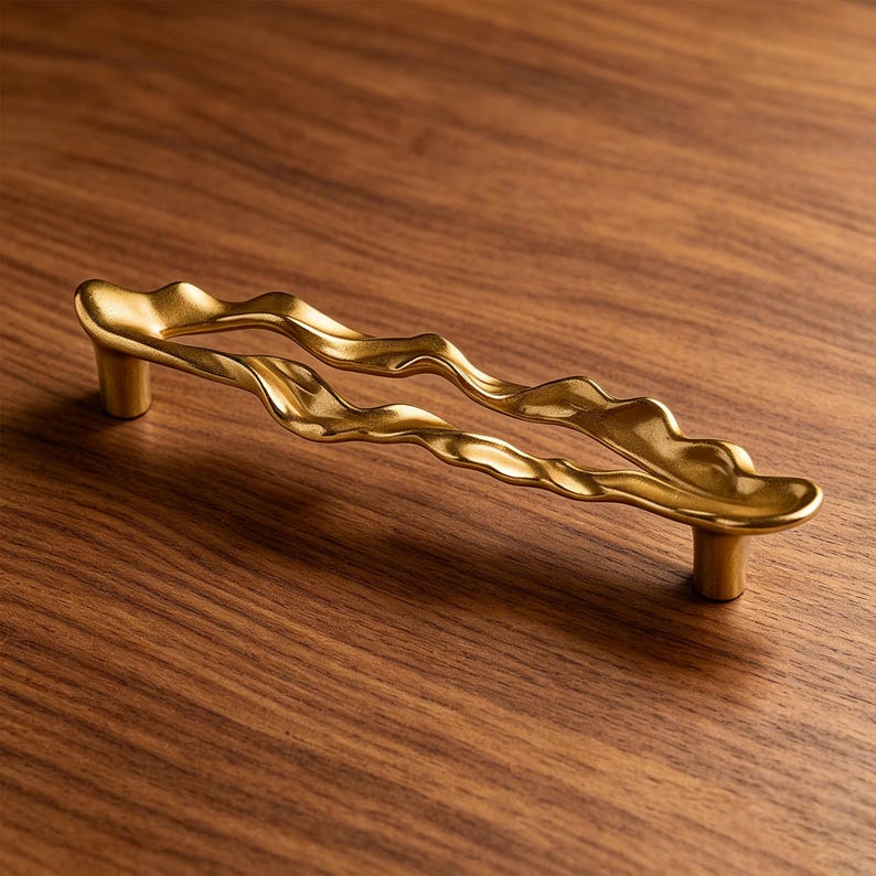 Puede incluir: Un tirador de mueble de metal dorado con una silueta larga, horizontal, ondulada y org&aacute;nica. El tirador est&aacute; sostenido por dos postes cil&iacute;ndricos en cada extremo, creando una forma continua, fluida y ondulante con un acabado suave.