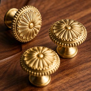 May include: Three circular brass cabinet knobs with a matte gold finish. Each knob features a central raised dot surrounded by a radial pattern of textured petals and a beaded outer edge, mounted on a tapered cylindrical base for attachment.
