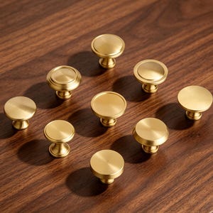 May include: A collection of eight circular cabinet knobs with a brushed gold finish. Each knob features a flat top surface and a tapered base, arranged in a scattered pattern. The hardware has a metallic sheen and a smooth, uniform texture.