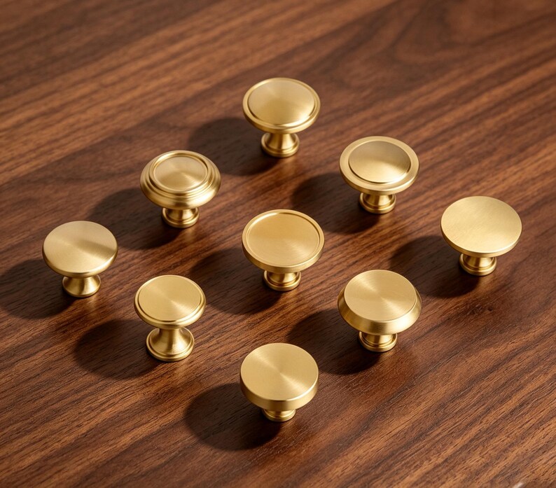 May include: A collection of eight circular cabinet knobs with a brushed gold finish. Each knob features a flat top surface and a tapered base, arranged in a scattered pattern. The hardware has a metallic sheen and a smooth, uniform texture.
