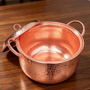 Puede incluir: Una olla redonda de cobre martillado con dos asas laterales y una tapa a juego. Las superficies exterior e interior presentan una textura con hoyuelos distintiva, reflejando un tono marrón anaranjado metálico en todo el recipiente.