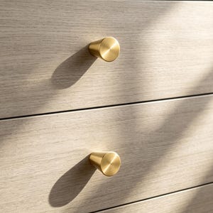 May include: A light-colored wooden dresser featuring two horizontal drawers, each equipped with a single, circular, brushed brass knob. The wood grain is visible across the drawer fronts, and the knobs cast distinct shadows on the surface.