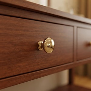 May include: A dark brown wooden drawer front featuring a smooth, polished, round gold-coloured metal knob centred on the panel. The wood grain is visible across the surface, and the drawer has a clean, rectangular frame design with a flat edge.