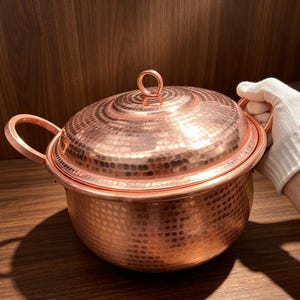 Op de afbeelding: Een ronde, gehamerde koperen pot met twee zijhandgrepen en een bijpassend deksel met een centrale ringgreep. Het buitenoppervlak van zowel de pot als het deksel vertoont een consistent, getextureerd gehamerd patroon met een glanzende afwerking.