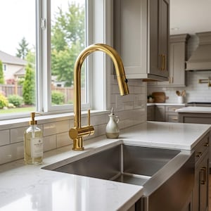 Può includere: Un rubinetto da cucina in oro lucido con design a collo di cigno ad arco alto e una singola leva laterale, posizionato sopra un lavello in acciaio inossidabile incassato in un piano di lavoro in marmo bianco con venature grigie.
