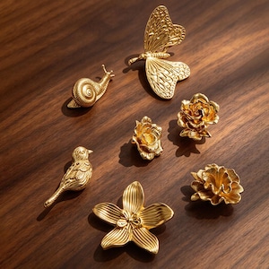 May include: A collection of seven gold-toned metal decorative pins or ornaments featuring a butterfly, a snail, a bird, and four distinct floral shapes. Each piece displays intricate embossed textures and a polished metallic finish throughout.